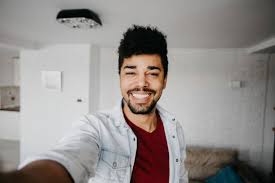Man with dark hair and beard wearing a red shirt and light denim jacket taking a selfie